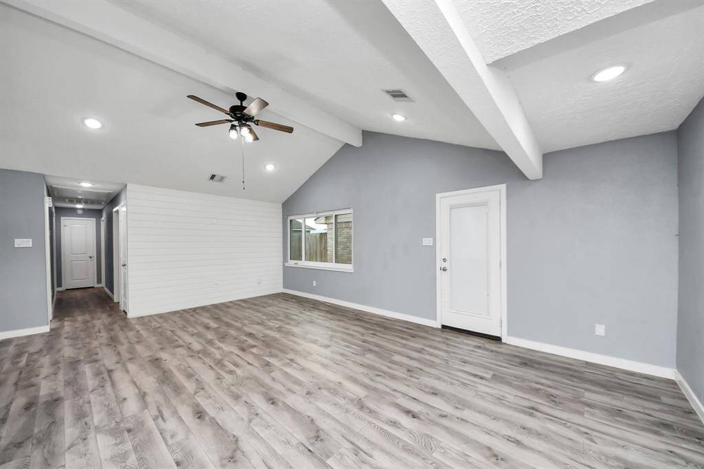 This spacious living area features vaulted ceilings with recessed lighting and a ceiling fan. The light gray walls and wood-style flooring create a modern, airy feel. A large window and glass door bring in natural light, leading to an outdoor area.