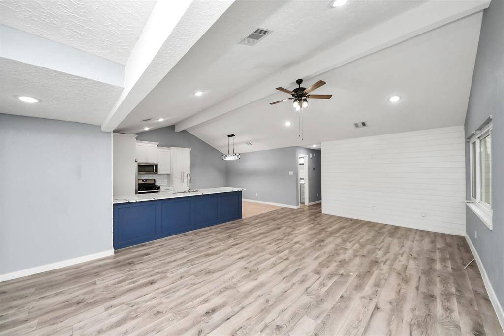 This spacious open-concept living area features elegant hardwood floors, a vaulted ceiling with a fan, and abundant natural light. The modern kitchen includes a large island with a blue base and white cabinetry, providing ample storage and a sleek look.
