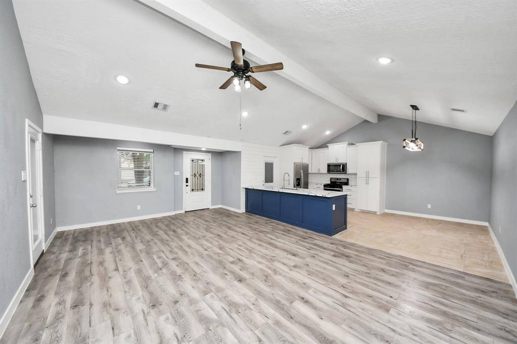 This open-concept living area features vaulted ceilings, modern gray flooring, and a spacious kitchen with a blue island and stainless steel appliances. Natural light enhances the space, creating a bright and welcoming environment.