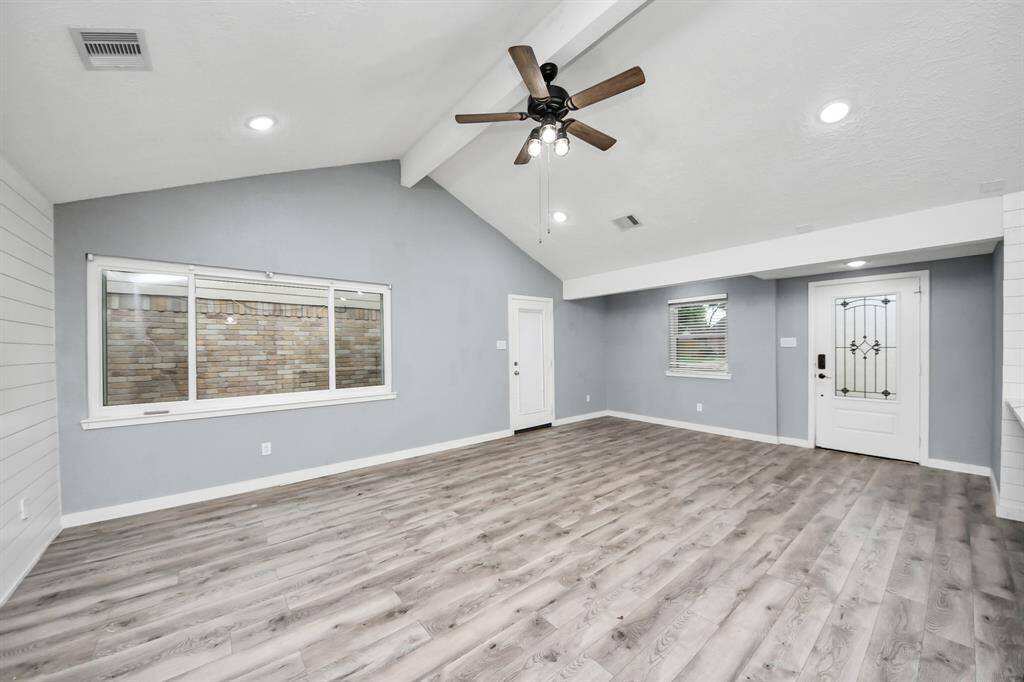 Bright and spacious living area featuring vaulted ceilings, a ceiling fan, modern recessed lighting, and large windows. The light gray wood flooring complements the soft blue-gray walls, creating a welcoming and airy atmosphere.
