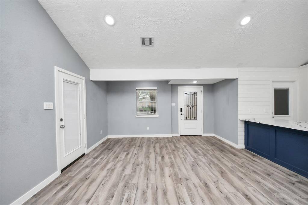 This room features modern light gray walls, sleek wood-style flooring, and recessed lighting. It includes a door with a decorative glass panel, a window for natural light, and a stylish white and blue bar area, creating an inviting and contemporary space.