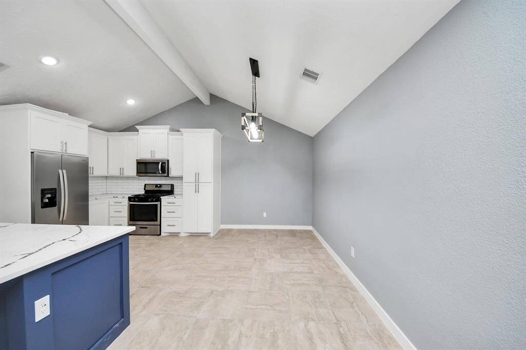 This photo showcases a modern kitchen and dining area with vaulted ceilings. It features stainless steel appliances, white cabinetry, a sleek blue island with a marble countertop, and soft gray walls. The space is well-lit, creating an inviting atmosphere.