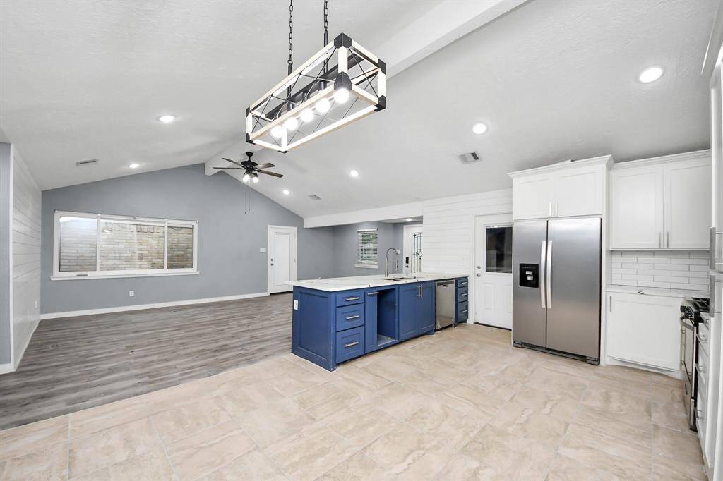 This spacious open-concept kitchen and living area features modern finishes, including a large island with blue cabinetry, stainless steel appliances, and recessed lighting. The room boasts a vaulted ceiling with a fan and a mix of tile and wood flooring, creating a bright and inviting atmosphere.