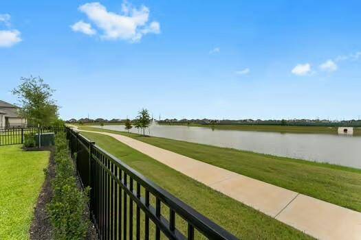 This photo showcases a backyard view with a black metal fence, overlooking a the lake and a paved walking path. The area is surrounded by grassy lawns and a few young trees, offering a peaceful and open outdoor space.