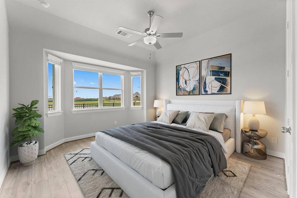 Primary Suite features large bay windows with lake views, a cozy bed with modern decor, a ceiling fan, and soft lighting, creating a tranquil and inviting atmosphere.