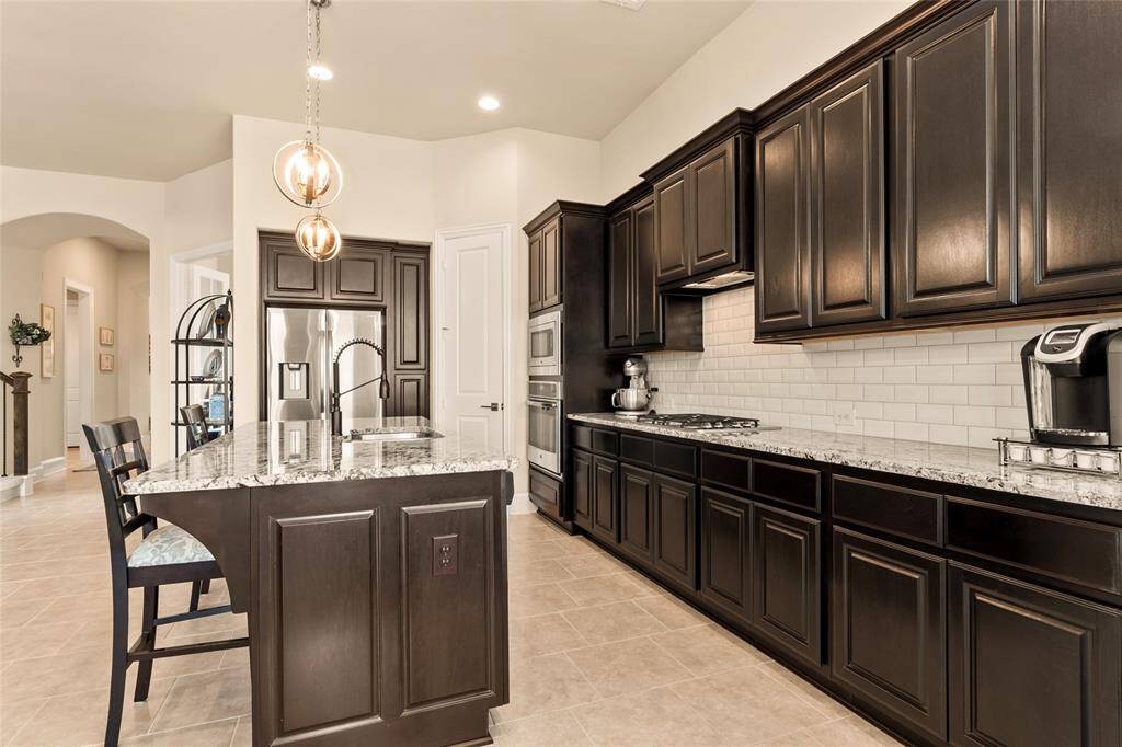 Pre-plumbed for water softener and Samsung kitchen refrigerator included, this kitchen features dark wood cabinetry with granite countertops and tile backsplash, and stainless steel appliances. It has a modern, open layout with pendant lighting and tiled flooring, offering ample space for cooking and dining.