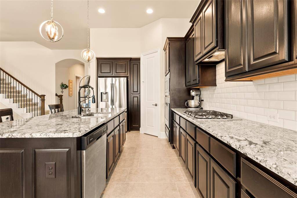 This kitchen features elegant dark wood cabinetry, granite countertops, and stainless steel appliances. It includes a spacious island with a modern sink and faucet, pendant lighting, and a gas cooktop. The open layout connects to the main living area.