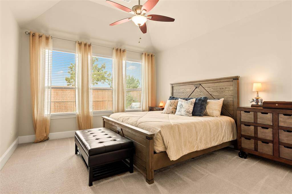 Primary bedroom with wall-of-windows, neutral paint tones, ceiling fan and sheer drapes creating a warm and comfortable space.