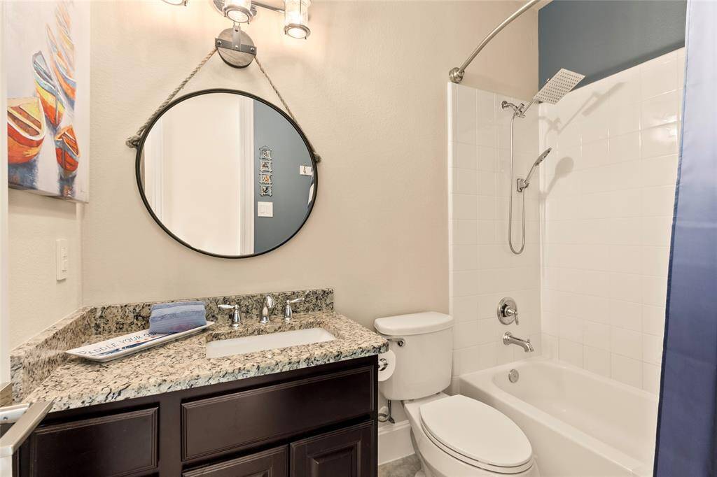 Gathroom features a modern design with a granite countertop, a round mirror, and stylish lighting. It includes a bathtub with a shower and a tiled surround, all in a neutral color scheme. A touch of color is added for a vibrant amibance.