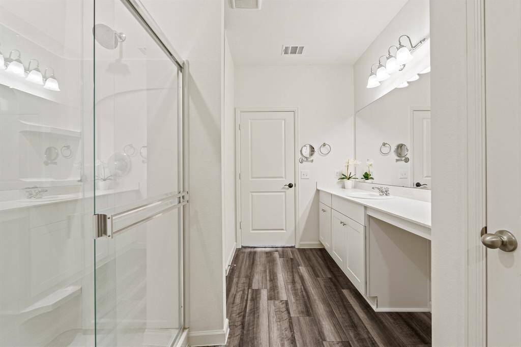 The Primary Bath boasts quartz countertops, a makeup vanity, wall-mounted magnifying glass, and roomy shower. The door to the linen closet can be seen to the right, providing the ideal spot to store additional towels and linens.