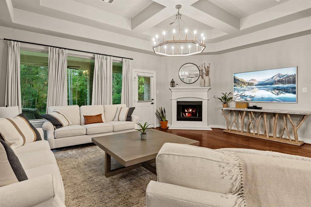This inviting open-concept living area features elegant coffered ceilings, hardwood floors, and large windows with lush green views.