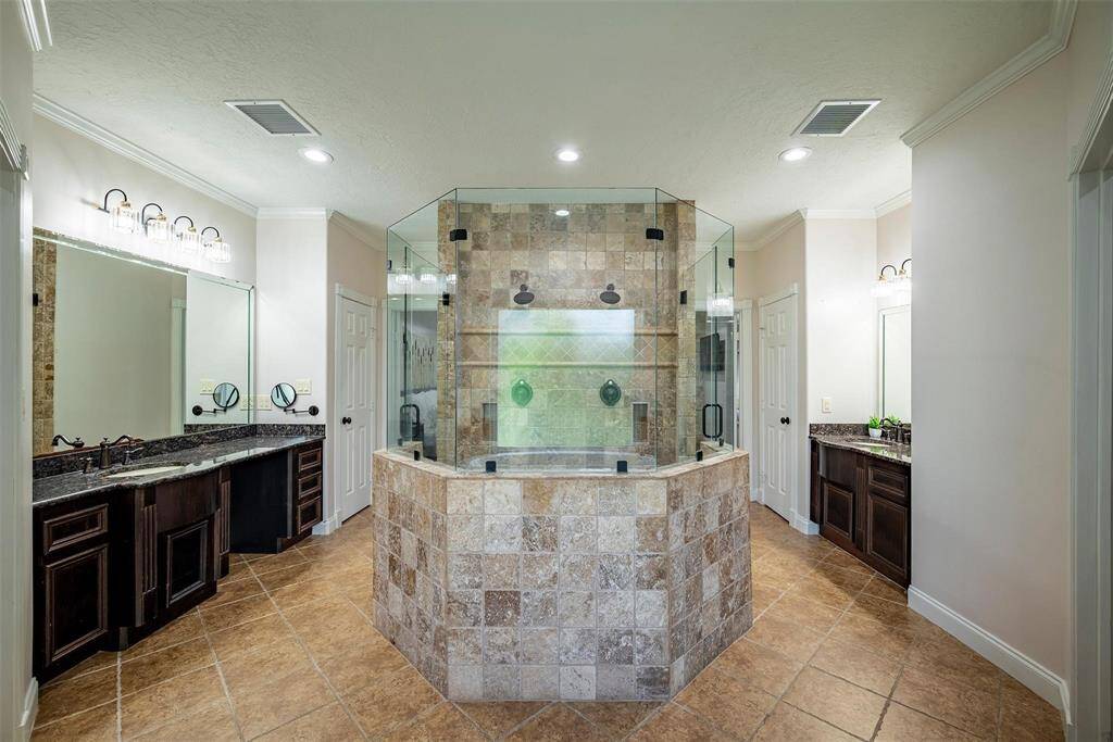 This luxurious bathroom features a central glass-enclosed shower with intricate tile work, dual vanities with granite countertops, and elegant lighting, creating a spa-like atmosphere.