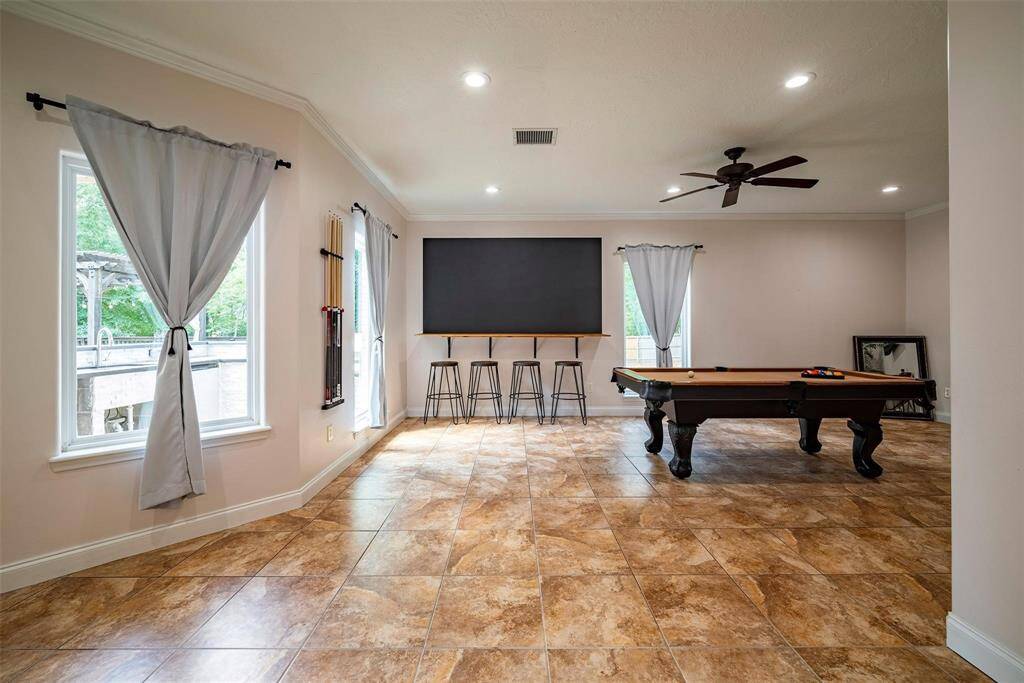 This spacious recreation room features tile flooring, a pool table, large windows with gray curtains, and a ceiling fan. It's perfect for entertaining, with ample natural light and a sleek, modern feel.
