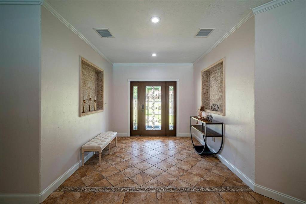 The photo shows a well-lit entryway featuring double glass doors with decorative panels. The space has a tiled floor with an elegant pattern, a cushioned bench, and a modern console table with decorative items. Recessed lighting adds a bright, welcoming ambiance.
