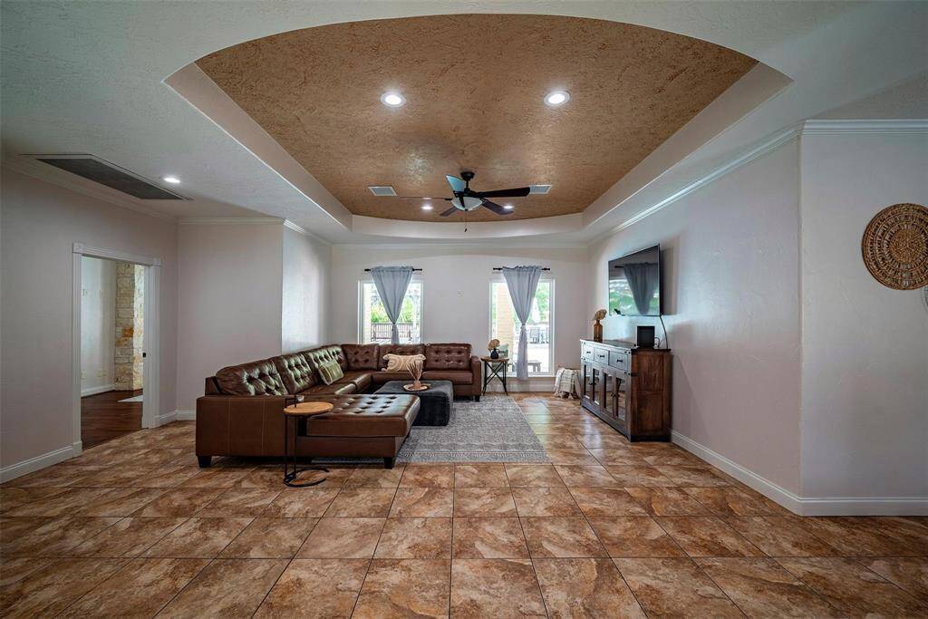 Spacious living room with a warm, earthy design featuring tiled floors and a coffered ceiling. It includes a large sectional sofa, a wall-mounted TV, and ample natural light from two windows.