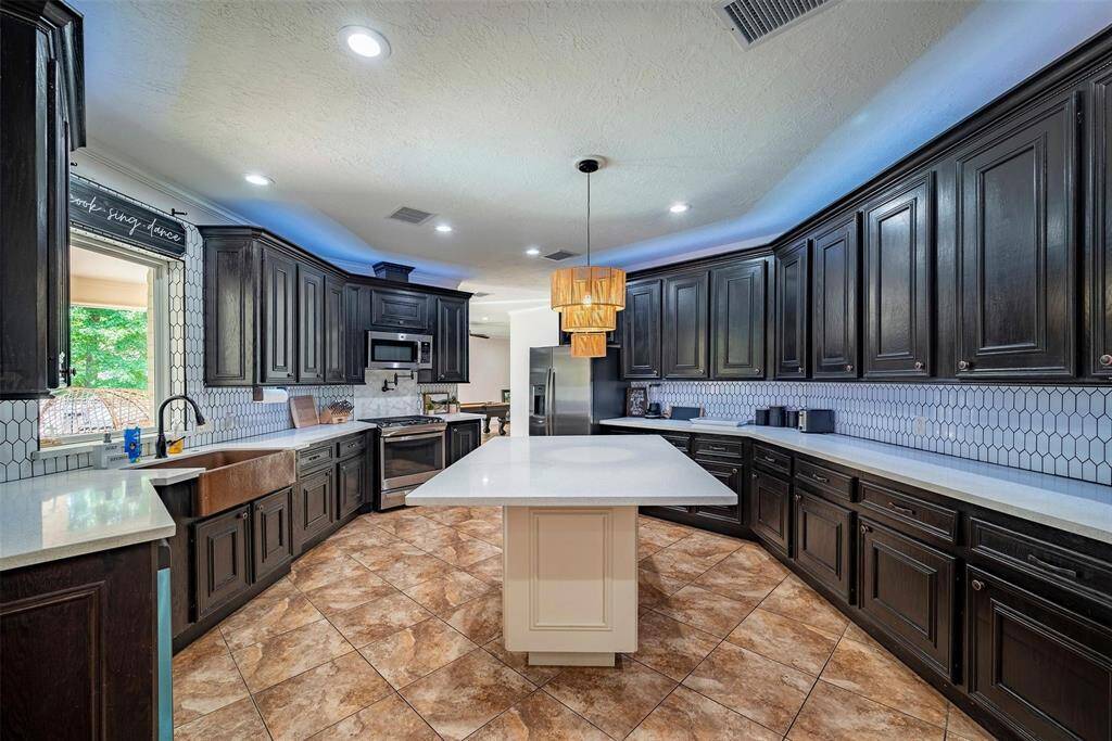 This kitchen features dark wood cabinetry, a central island with a modern pendant light, and a stainless steel appliance suite. The white countertops and hexagonal tile backsplash contrast beautifully with the warm, tiled floor. Ample natural light streams through a window over the sink, creating an inviting and functional space.