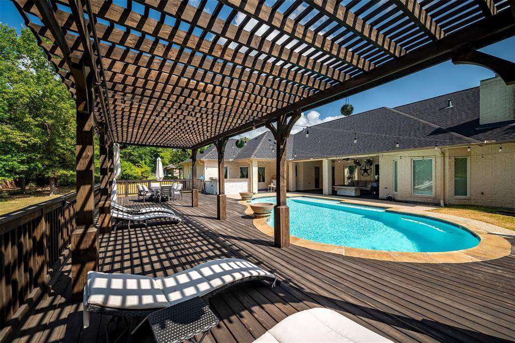 A backyard with a spacious wooden deck and pergola, featuring lounge chairs and a dining area. It overlooks a large, inviting pool adjacent to a modern house. Ideal for outdoor relaxation and entertainment.
