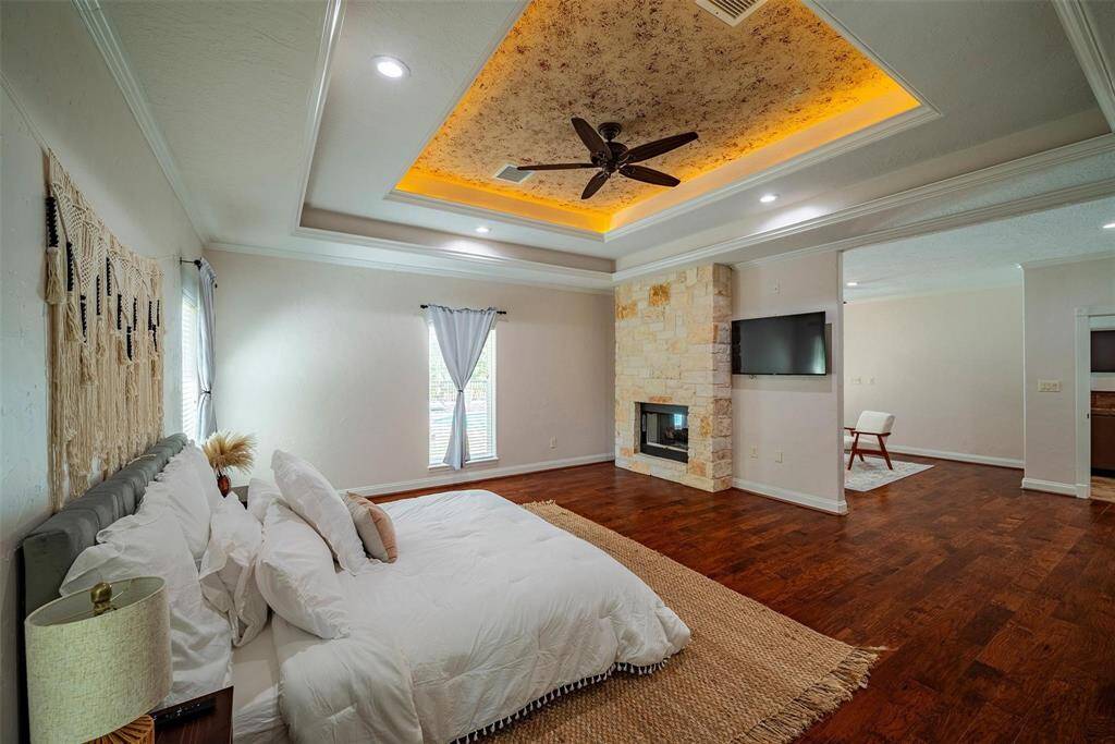 This spacious bedroom features a cozy fireplace, elegant hardwood floors, and a decorative tray ceiling with recessed lighting and a ceiling fan. Natural light fills the room through two large windows, complementing the serene, neutral decor. Perfect for relaxation and comfort.
