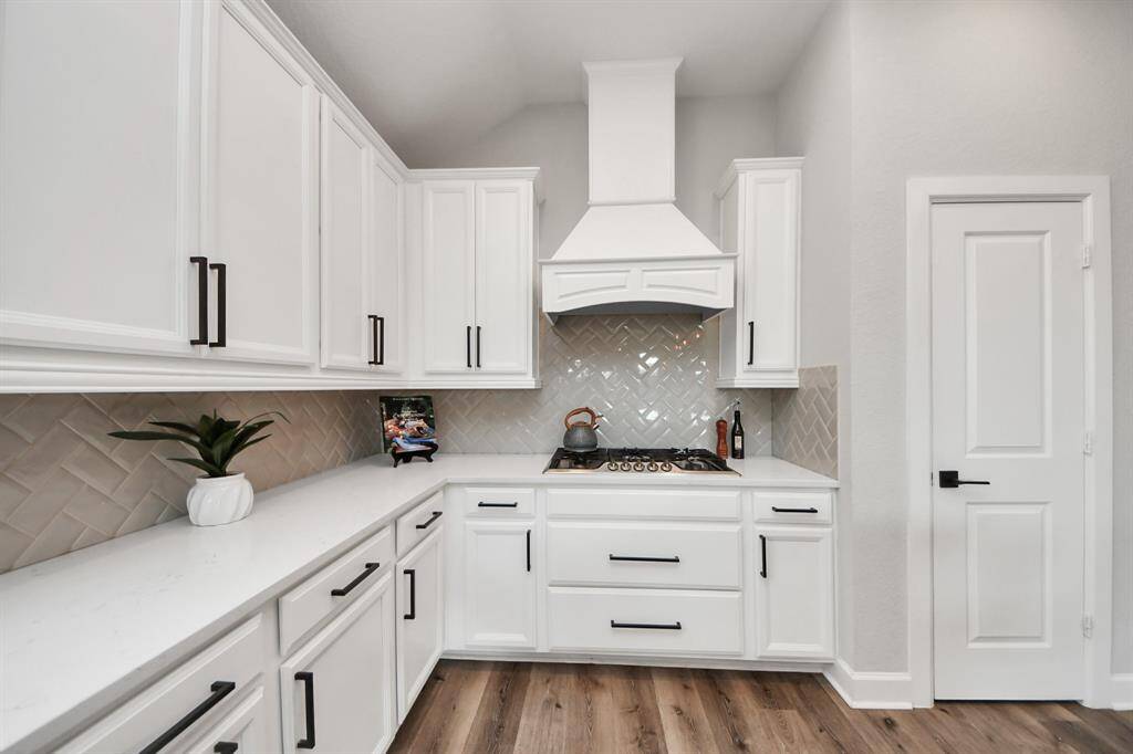 This kitchen features elegant white cabinetry with sleek black handles, a modern gas stovetop with a stylish hood, and a chic herringbone tile backsplash, and complemented by ample counter space and a clean, bright aesthetic.
