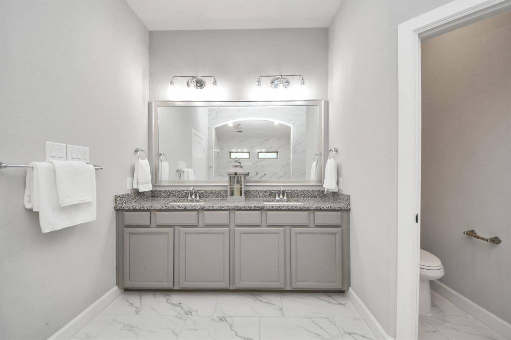 This bathroom features a sleek design with a large granite double vanity, ample storage, and a spacious mirror. The room has a bright, clean aesthetic with light gray walls and marble tile flooring. A walk-in shower with a rainfall showerhead.