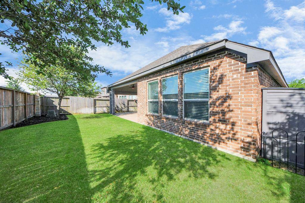 A well-maintained backyard with green lawn, mature trees, and a wooden fence. The brick exterior of the house features large windows, providing a bright and inviting atmosphere.