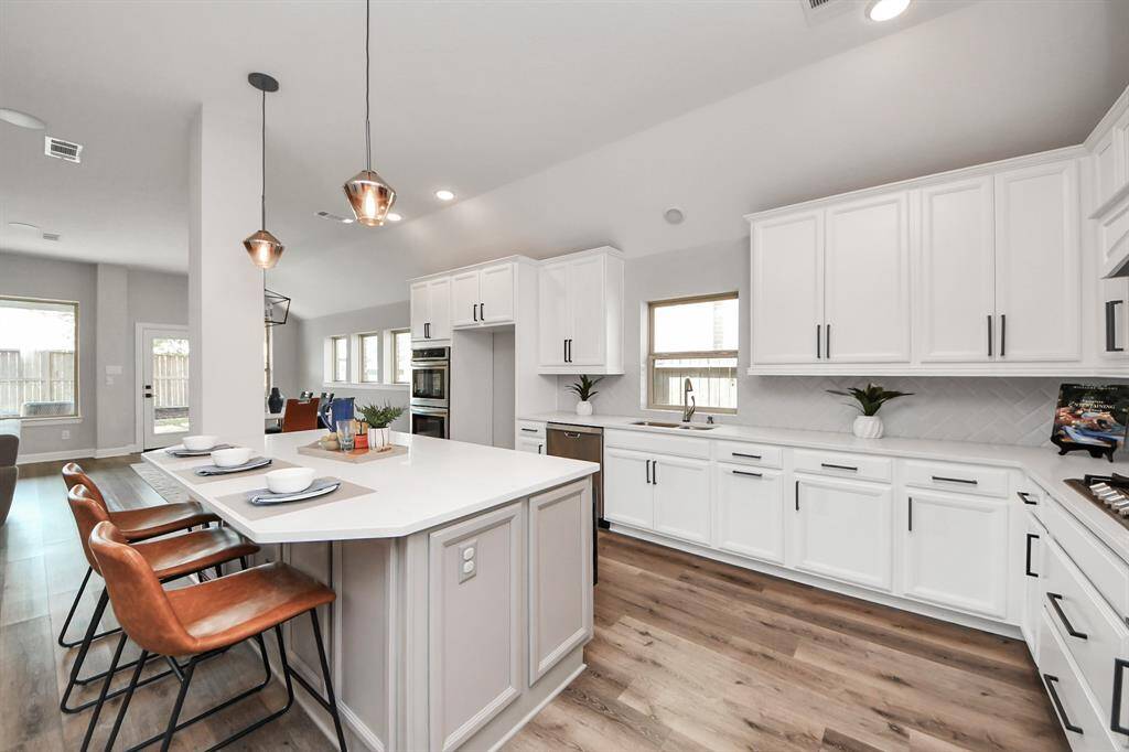 Kitchen features a spacious, open layout with sleek white cabinetry, a large island with seating, and stainless steel appliances. Pendant lighting adds a stylish touch, while the wood flooring complements the contemporary design. Perfect for entertaining and family gatherings.