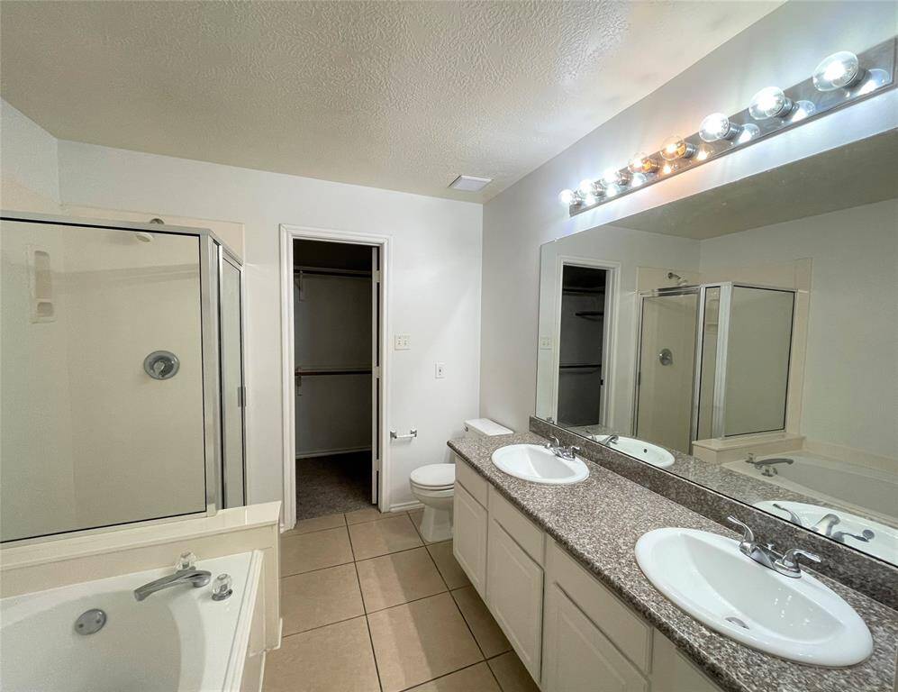 Bright bathroom with dual sinks, granite countertops, a walk-in shower, and a separate bathtub. There's ample lighting above the mirror and a door leading to a walk-in closet.