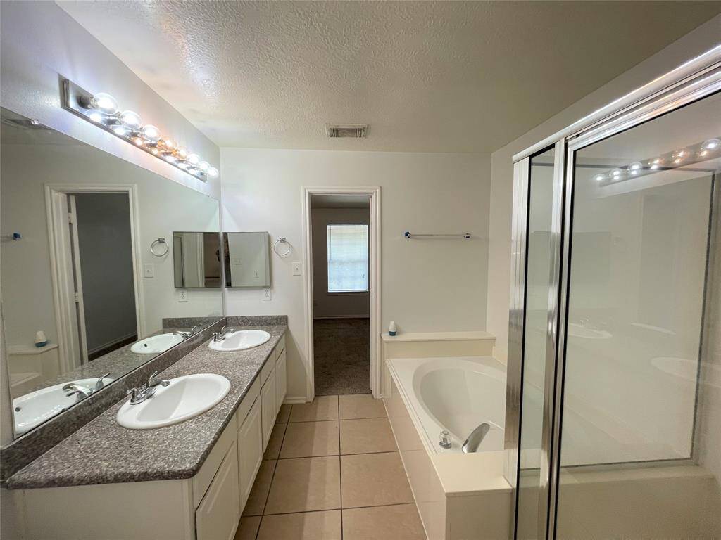 This bathroom features a double sink vanity with granite countertops, a large mirror with overhead lighting, a spacious bathtub, and a glass-enclosed shower. The neutral tile flooring and soft wall colors create a clean, modern look.