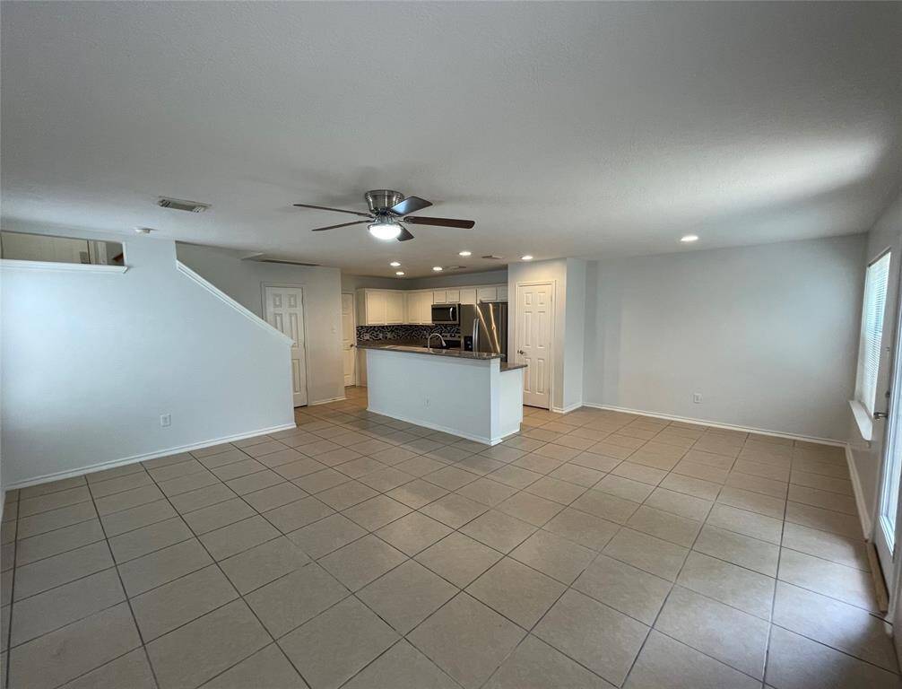Spacious open-concept living area with tiled floors, modern ceiling fan, and ample natural light. The kitchen features stainless steel appliances and a stylish backsplash. Stairs lead to the upper level, enhancing the home's layout.