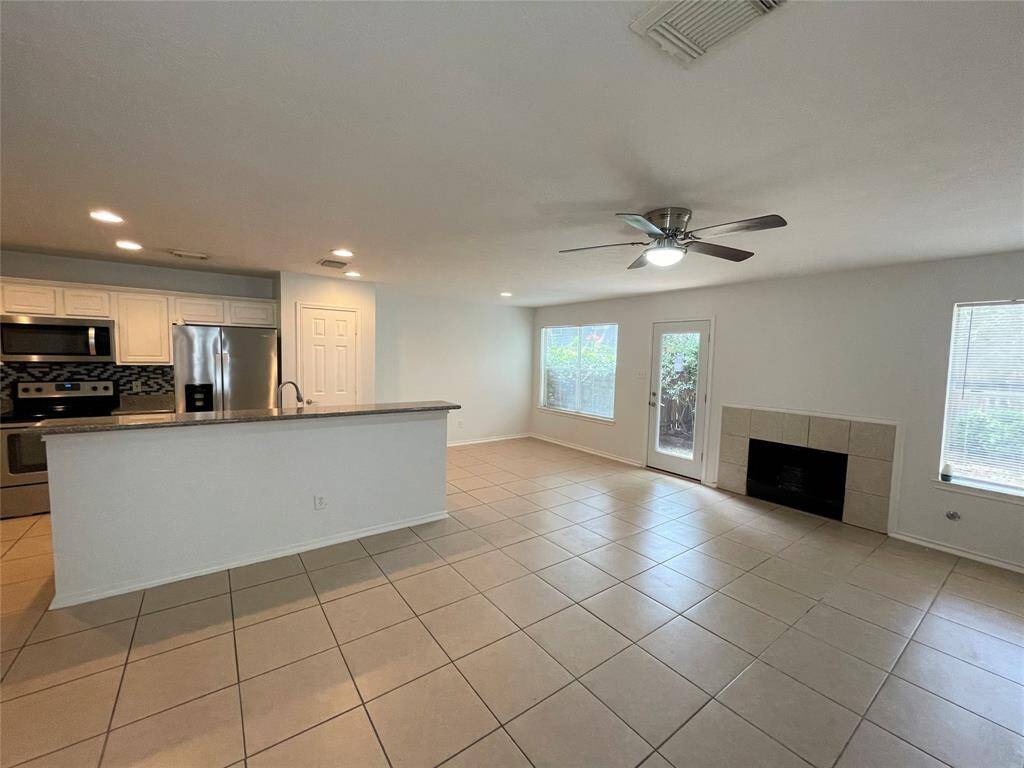 This spacious, open-concept living area features a modern kitchen with stainless steel appliances and a stylish backsplash. It has tile flooring, a cozy fireplace, and ample natural light from large windows and a glass door leading outside, creating a welcoming and functional space.