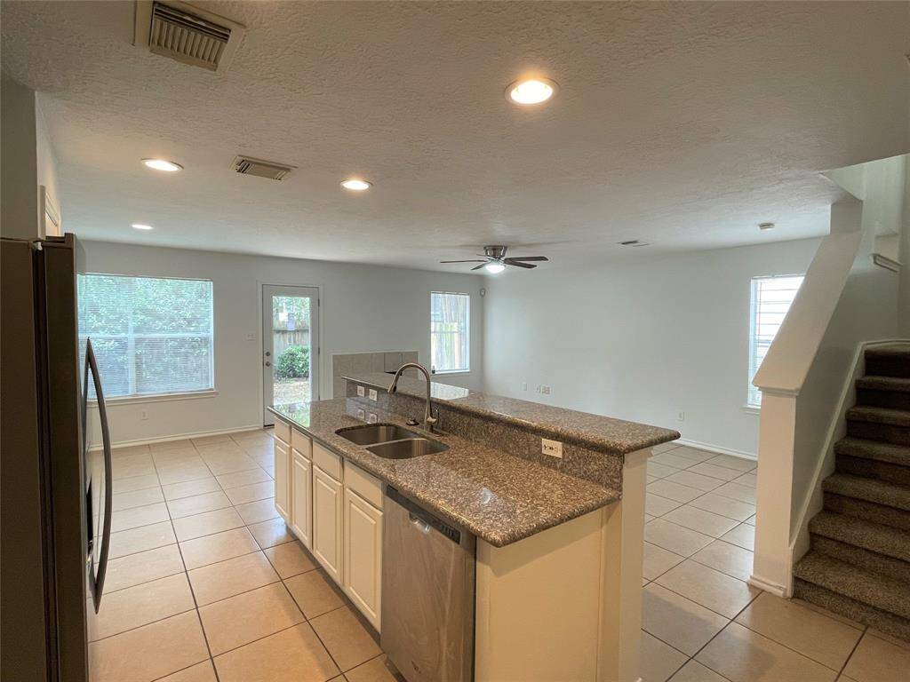 spacious, open-concept kitchen and living area with tile flooring. The kitchen features granite countertops, a double sink, and white cabinetry. There is ample natural light from large windows, and the room includes recessed lighting and a ceiling fan. A staircase leads to the upper level.