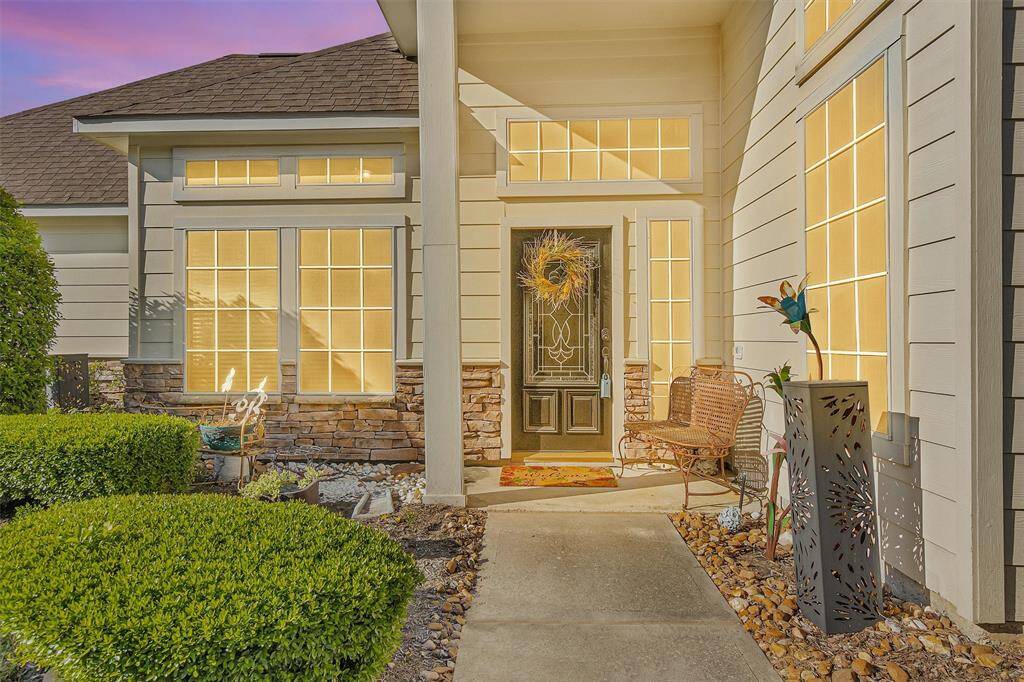 This inviting home entrance features a stylish front door with decorative glass, surrounded by large, bright windows. The exterior combines stone and siding, with well-maintained landscaping and a cozy seating area, creating a warm, welcoming atmosphere.