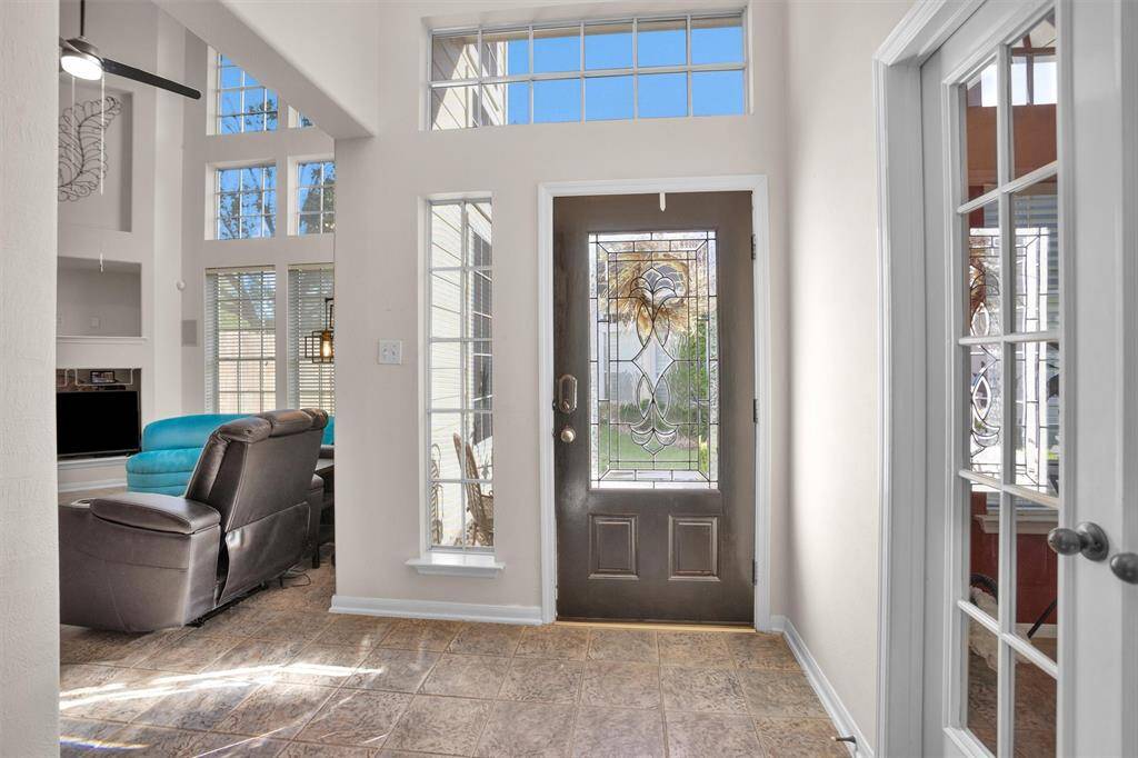 Bright entryway with decorative glass door, tiled floor, and high ceilings. Adjacent living area features large windows and a cozy seating arrangement.