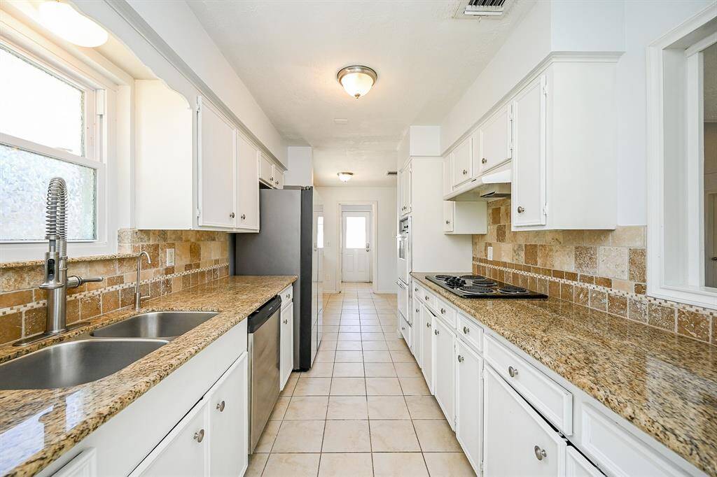 Bright galley kitchen with granite countertops, white cabinetry, and stainless steel appliances. It features a double sink, tiled backsplash, and ample storage, offering a functional and stylish cooking space.