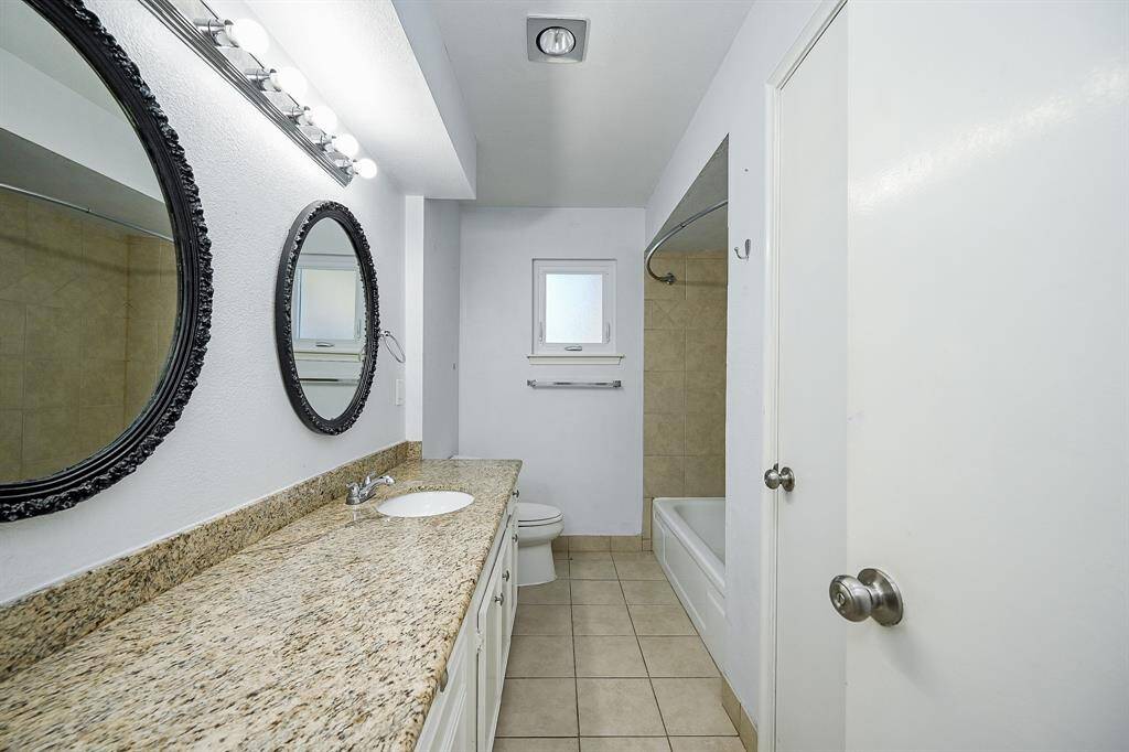 This bathroom features a long granite countertop with a single sink, framed mirrors, a tiled floor, and a shower-tub combo. It has a bright, clean look with ample lighting and a small window for natural light.