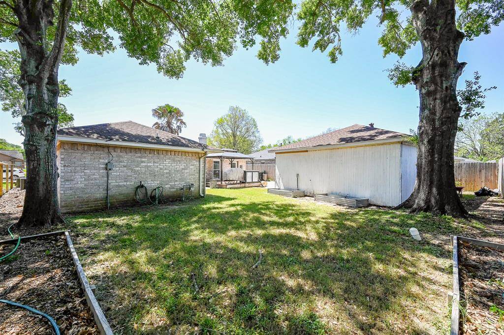 This photo showcases a spacious backyard with mature trees providing shade, bordered by two outbuildings. It offers ample space for outdoor activities or gardening, with a private, fenced perimeter.
