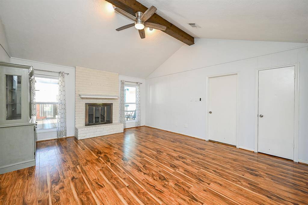 This spacious family room features vaulted ceilings with a wooden beam, and a cozy brick fireplace. The hardwood floors add warmth and elegance, while neutral walls offer a versatile backdrop for decorating.