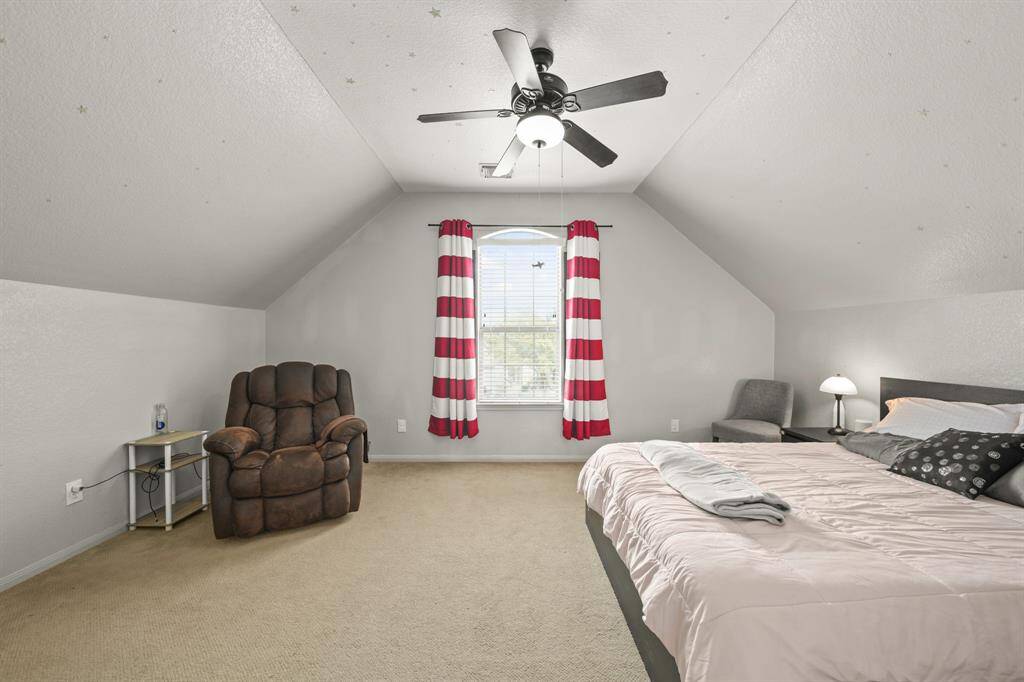 Large secondary bedroom with carpet floors, a large window with privacy blinds, and a vaulted ceiling with a ceiling fan.