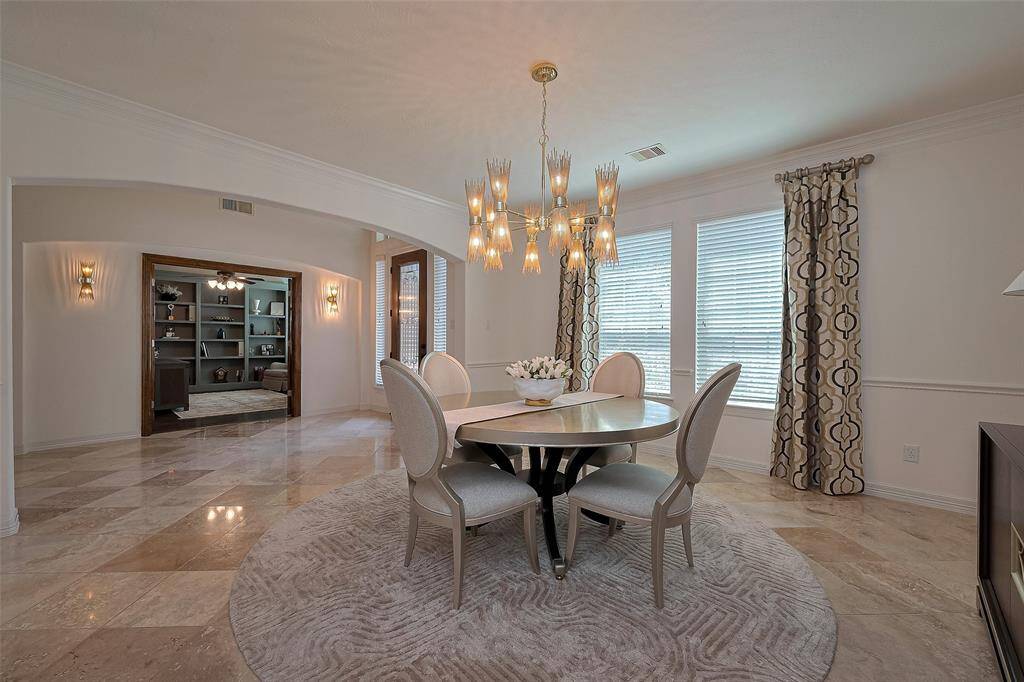 This elegant dining room features a round table with stylish chairs, a modern chandelier, and large windows with patterned curtains. The space has tile floors and opens to the foyer, adding a touch of sophistication and functionality.