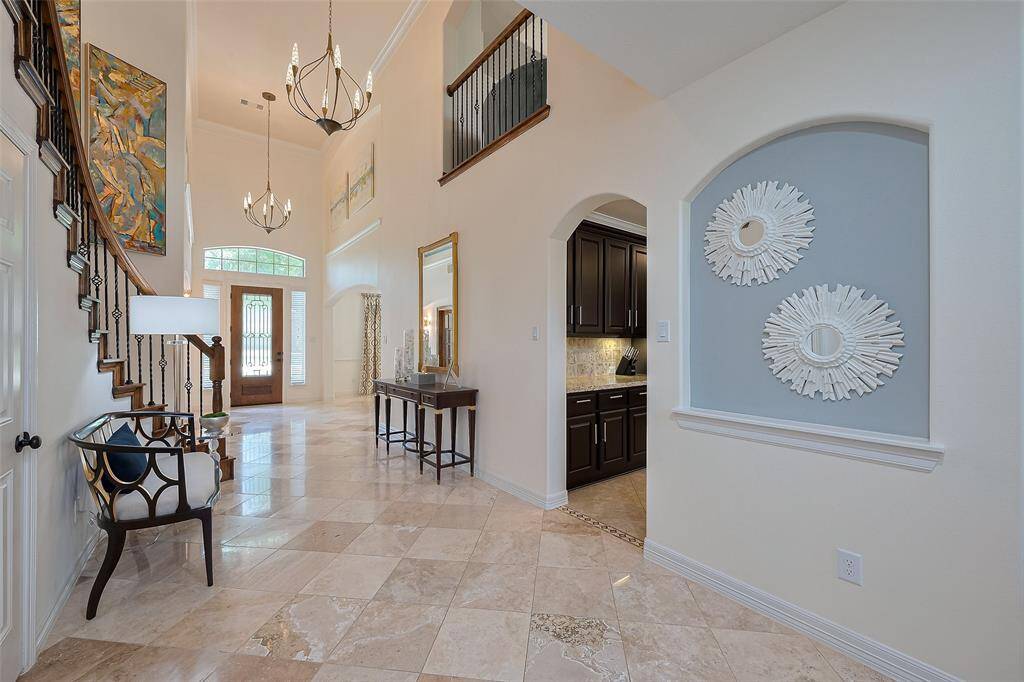 This elegant foyer features high ceilings, a grand staircase, and polished tile flooring. Natural light streams through large windows by the front door, highlighting the stylish decor and leading into a kitchen area with dark cabinetry. The newly installed chandelier adds a touch of sophistication, creating a welcoming entryway.