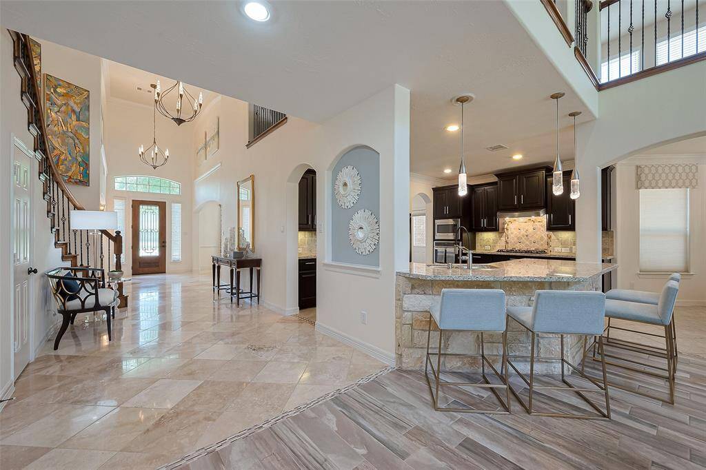 This spacious entryway features elegant tile flooring, high ceilings, and a grand staircase. The open kitchen boasts a breakfast bar, modern lighting, and dark cabinetry, creating a welcoming and stylish atmosphere.