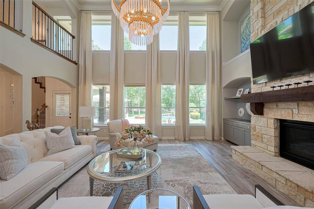 This spacious family room features high ceilings and large windows with new custom curtains, allowing for abundant natural light. A stone fireplace and elegant chandelier by John Richards add warmth and sophistication. The open layout is perfect for entertaining, with stylish furnishings and a balcony view from above.