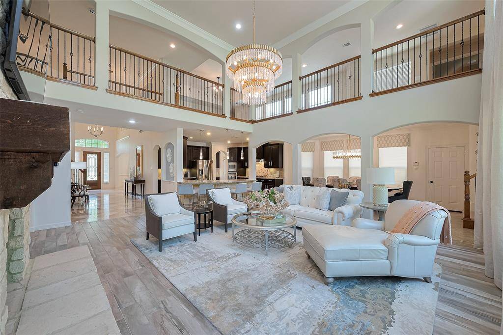 This photo showcases a spacious, elegantly designed open living area with high ceilings and a grand chandelier by John Richards. The room features a cozy seating arrangement, modern kitchen with a breakfast bar, and a view of the staircase and upper catwalk. The freshly painted neutral color palette and ample natural light enhance its inviting atmosphere.