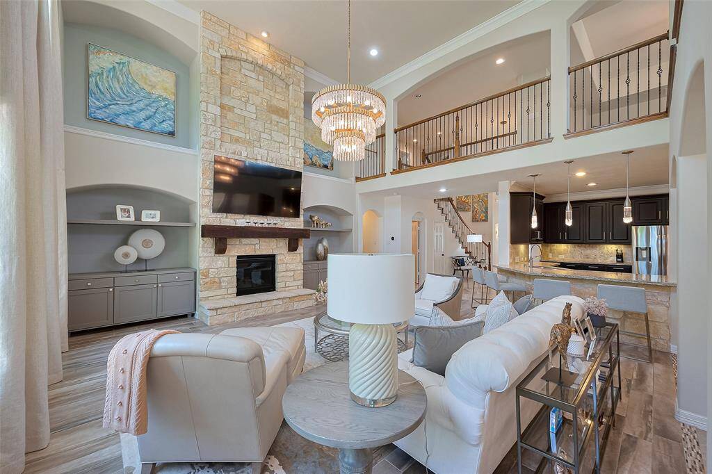This spacious living area features high ceilings and an open floor plan. It includes a stone fireplace, elegant chandelier, and large windows for natural light. The kitchen boasts modern appliances and a breakfast bar, while the mezzanine adds an extra touch of sophistication.