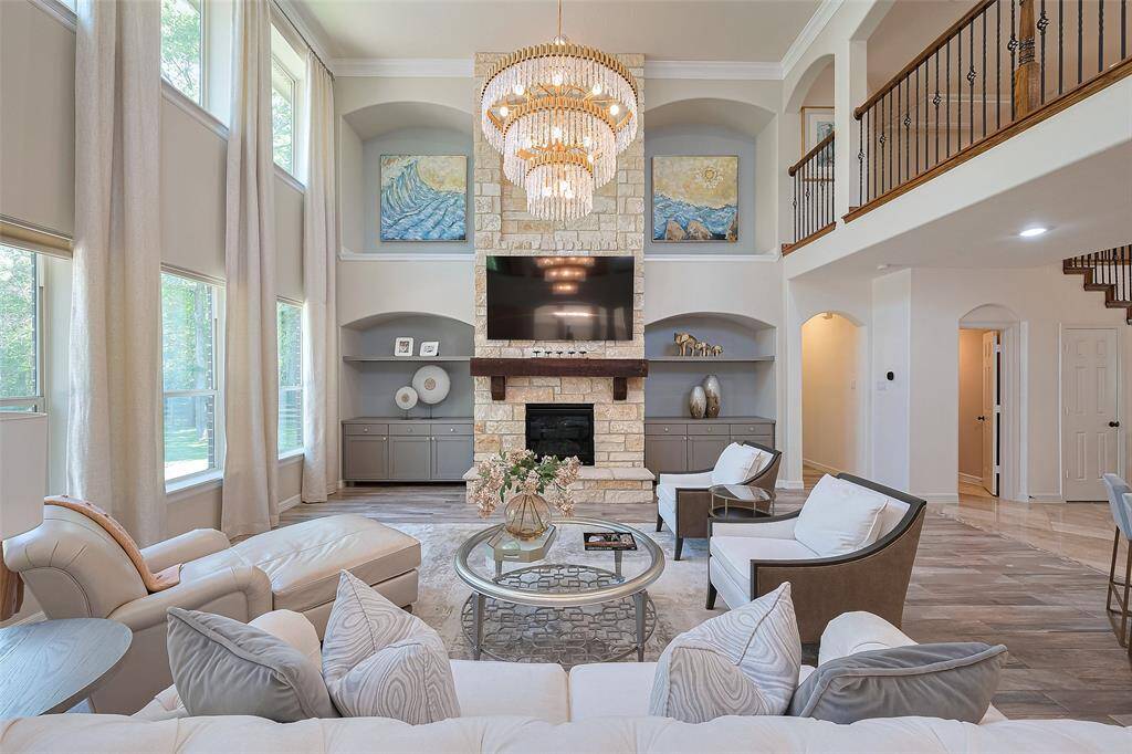 This spacious family room features high ceilings, large windows with custom dressings, and a stone fireplace with a mounted TV. Elegant chandeliers and neutral-toned furniture create a warm, inviting atmosphere. There's a blend of modern and classic design elements, complemented by a visible staircase and upper balcony area.