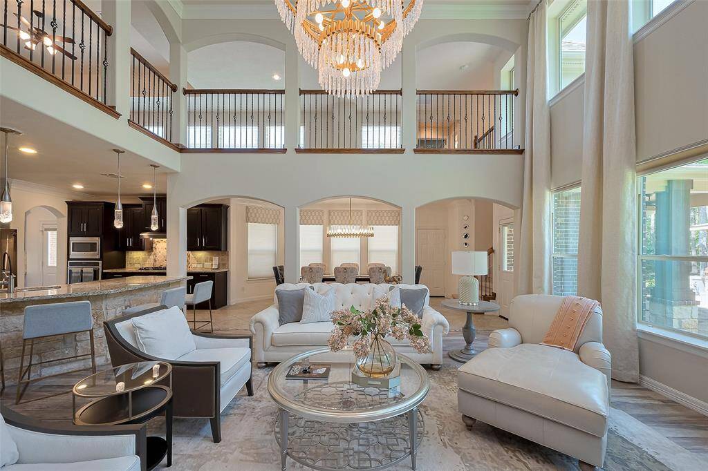 This elegant open-concept family room area features a high ceiling and large windows with custom curtains that fill the space with natural light. A grand chandelier adds sophistication, while the upstairs balcony overlooks the cozy seating area. The adjacent kitchen boasts modern fixtures, dark cabinetry, and a breakfast bar, perfect for entertaining.