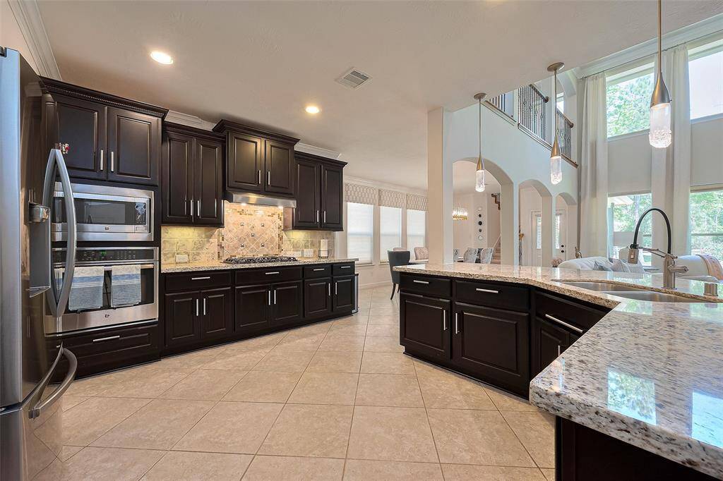 This modern kitchen features dark wood cabinetry, granite countertops, and stainless steel appliances. The open layout flows into a spacious living area with large windows and high ceilings, offering abundant natural light.