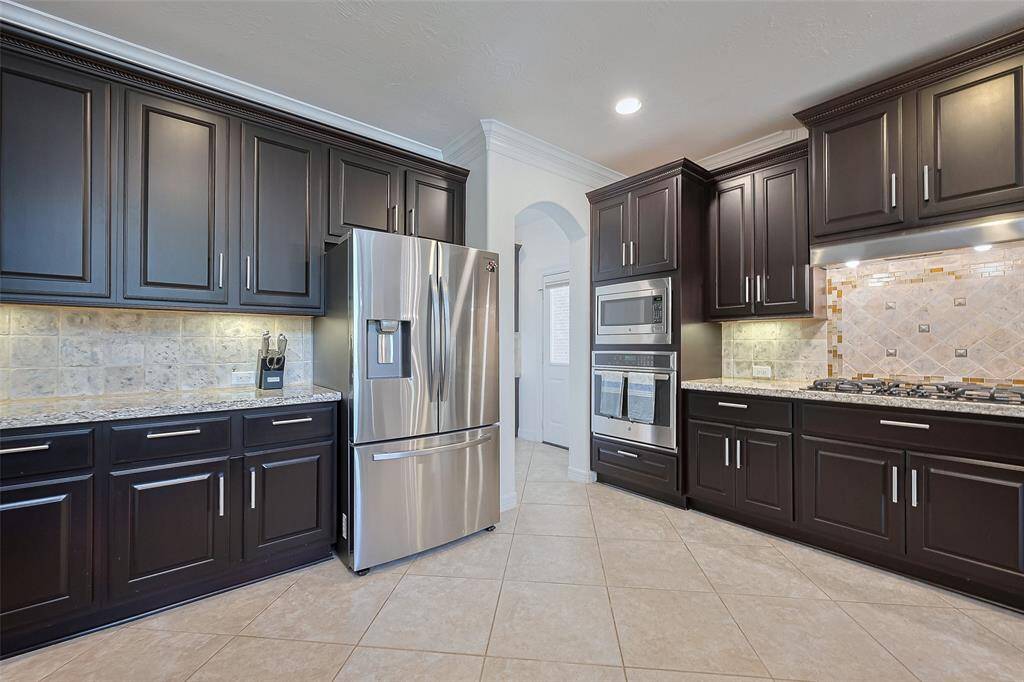This kitchen features sleek dark cabinetry, stainless steel appliances, and a stylish tile backsplash. It includes a spacious fridge, stacked microwave and oven, and a gas cooktop, all complemented by granite countertops and under-cabinet lighting.