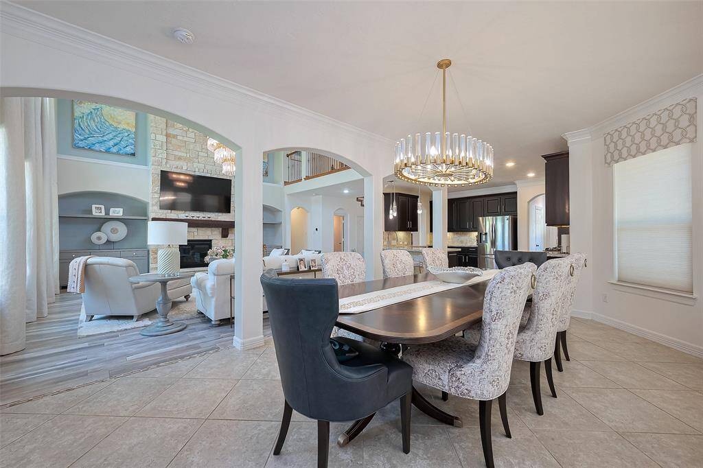 This spacious and elegant open-concept breakfast and family area features a modern chandelier, a cozy fireplace, and a sleek kitchen with dark cabinetry. The room is filled with natural light, creating a warm and inviting atmosphere, perfect for entertaining.