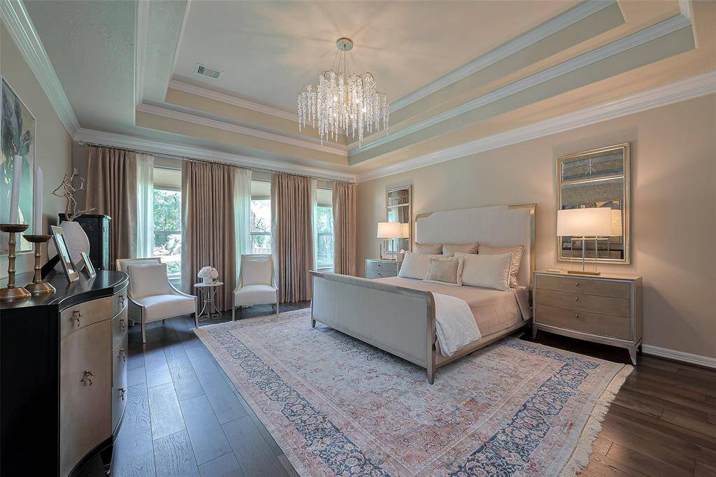 This elegant Primary bedroom features a tray ceiling with a stunning chandelier, large windows with new custom drapes, and new hardwood floors. The space includes a cozy bed, stylish nightstands, and seating, creating a luxurious and inviting atmosphere.